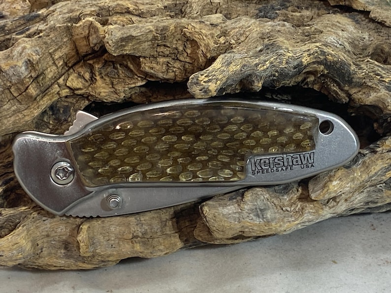 May include: A closed folding pocket knife with a metallic silver handle and a translucent amber-colored honeycomb patterned inlay. The handle features the text Kershaw SpeedSafe USA and a small circular hole at the end for a lanyard attachment.
