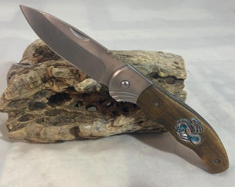 Könnte beinhalten: Ein silbernes Klappmesser mit einem Holzgriff, der mit einem blauen und silbernen Drachenmotiv verziert ist. Das Messer hat eine teilweise geöffnete Klinge.