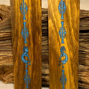 Könnte beinhalten: Zwei rechteckige Holzstücke mit einem Kokopelli-Design, das mit blauem Stein eingelegt ist. Das Holz hat einen warmen Braunton mit sichtbarer Maserung. Die Kokopelli-Figur wird beim Flötespielen dargestellt.