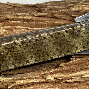 Pu&ograve; includere: Primo piano di un coltello tascabile con un manico con un motivo a scaglie nei toni del verde e del marrone. La lama &egrave; parzialmente visibile e il coltello ha una ghiera color argento e una piccola estensione del manico curva.