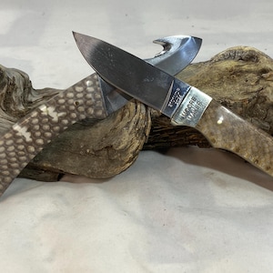 K&ouml;nnte beinhalten: Ein Klappmesser mit einer silberfarbenen Metallklinge und einem Aufrei&szlig;haken. Der Griff hat eine strukturierte, helle Oberfl&auml;che mit einem Muster und kleinen Metallstiften. Die Klinge ist mit dem Text Moore Maker 2024 graviert.