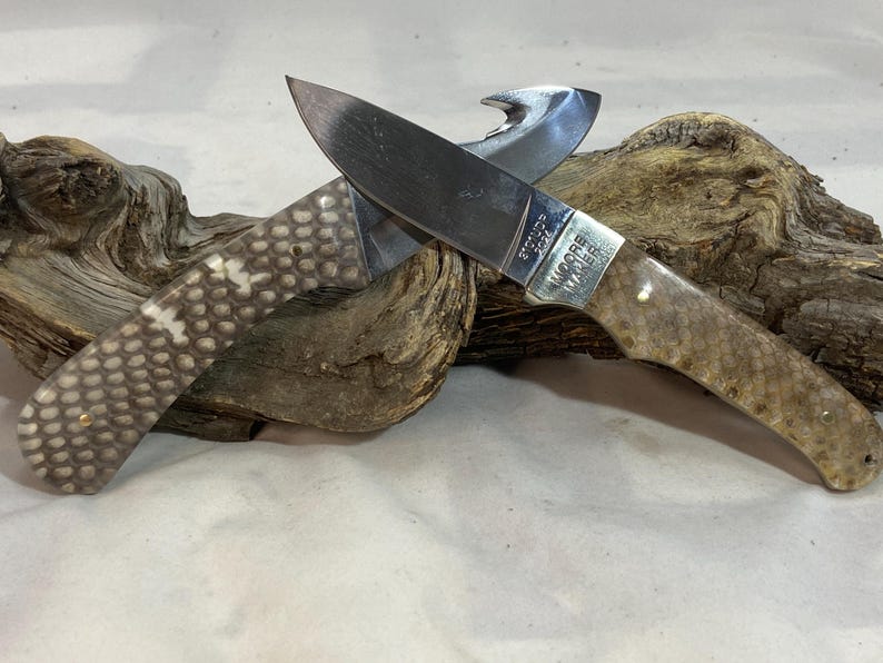 K&ouml;nnte beinhalten: Ein Klappmesser mit einer silberfarbenen Metallklinge und einem Aufrei&szlig;haken. Der Griff hat eine strukturierte, helle Oberfl&auml;che mit einem Muster und kleinen Metallstiften. Die Klinge ist mit dem Text Moore Maker 2024 graviert.