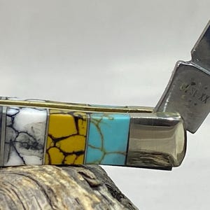 Puede incluir: Primer plano de una navaja de bolsillo con un mango decorativo. El mango presenta un dise&ntilde;o de mosaico con secciones negras, blancas, amarillas y turquesas. La hoja est&aacute; parcialmente abierta, revelando un acabado plateado y texto estampado.