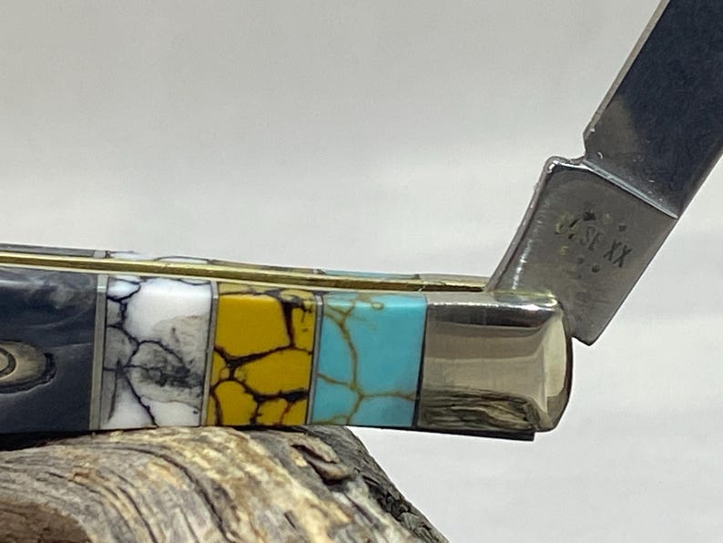 Puede incluir: Primer plano de una navaja de bolsillo con un mango decorativo. El mango presenta un dise&ntilde;o de mosaico con secciones negras, blancas, amarillas y turquesas. La hoja est&aacute; parcialmente abierta, revelando un acabado plateado y texto estampado.