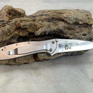 May include: A silver-toned folding pocket knife with a metallic handle featuring the Kershaw brand name and a pocket clip. The stainless steel blade is partially extended and displays the number 1880 along with a small, detailed snake graphic.