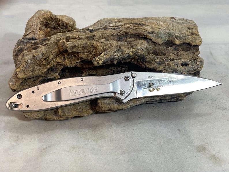 May include: A silver-toned folding pocket knife with a metallic handle featuring the Kershaw brand name and a pocket clip. The stainless steel blade is partially extended and displays the number 1880 along with a small, detailed snake graphic.