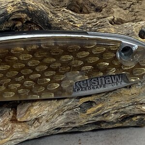 May include: A folding knife handle featuring a translucent amber-colored overlay with a repeating oval pattern. The metallic base displays the text Kershaw SPEEDSAFE USA in black lettering near a circular pivot hole on the right side.