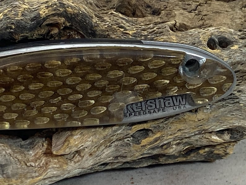 May include: A folding knife handle featuring a translucent amber-colored overlay with a repeating oval pattern. The metallic base displays the text Kershaw SPEEDSAFE USA in black lettering near a circular pivot hole on the right side.