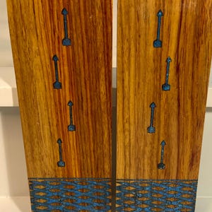 Könnte beinhalten: Zwei Holzpaneele mit warmer, rötlich-brauner Maserung. Jedes Paneel weist blaue Pfeileinlagen und ein blaues geometrisches Muster am Boden auf. Die Paneele werden nebeneinander dargestellt und zeigen die detaillierte Handwerkskunst.