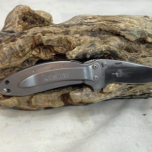 May include: A silver-toned folding pocket knife with a metallic handle featuring the Kershaw brand name. The blade is partially extended and displays the model number 1620FL along with a small etched graphic of a snake on the metal surface.