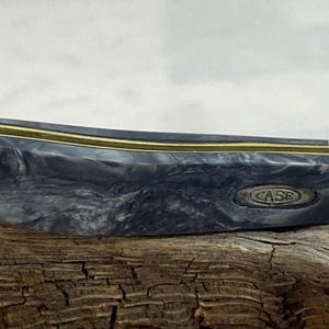 Puede incluir: Primer plano de una navaja de bolsillo con un mango gris y negro veteado. El cuchillo tiene un acento color lat&oacute;n y la palabra "CASE" est&aacute; grabada en el mango. El cuchillo est&aacute; sobre un trozo de madera.