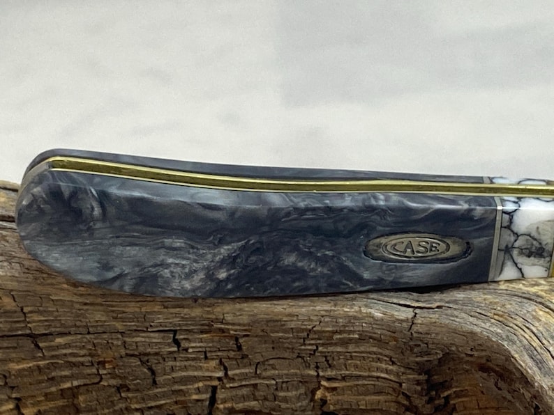 Puede incluir: Primer plano de una navaja de bolsillo con un mango gris y negro veteado. El cuchillo tiene un acento color lat&oacute;n y la palabra "CASE" est&aacute; grabada en el mango. El cuchillo est&aacute; sobre un trozo de madera.