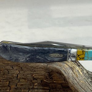 Puede incluir: Una navaja de bolsillo cerrada con un mango gris veteado y una hoja de acero inoxidable. El mango presenta una incrustaci&oacute;n decorativa de segmentos blancos, amarillos y turquesas. La palabra "CASE" es visible en el mango.