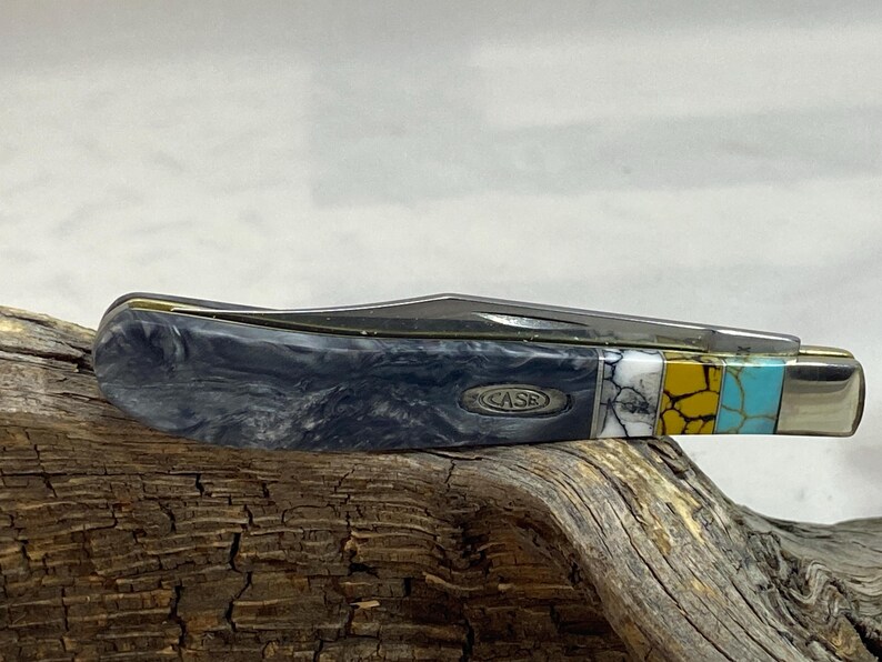 Puede incluir: Una navaja de bolsillo cerrada con un mango gris veteado y una hoja de acero inoxidable. El mango presenta una incrustaci&oacute;n decorativa de segmentos blancos, amarillos y turquesas. La palabra "CASE" es visible en el mango.