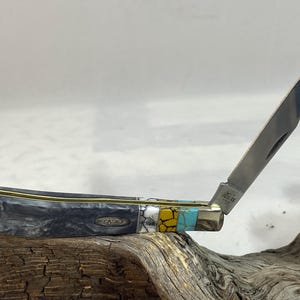 Puede incluir: Una navaja de bolsillo Case con una hoja parcialmente abierta. El mango es una mezcla de material veteado negro y gris, acentuado con ribetes dorados y secciones turquesa, amarillas y blancas. La hoja es plateada.