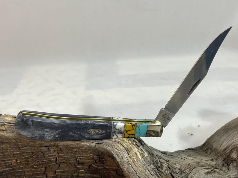 Puede incluir: Una navaja de bolsillo Case con una hoja parcialmente abierta. El mango es una mezcla de material veteado negro y gris, acentuado con ribetes dorados y secciones turquesa, amarillas y blancas. La hoja es plateada.