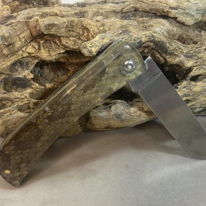 May include: A folding pocket knife with a partially open, stainless steel blade. The handle has a mottled green and brown pattern. The knife is resting on a piece of weathered wood.