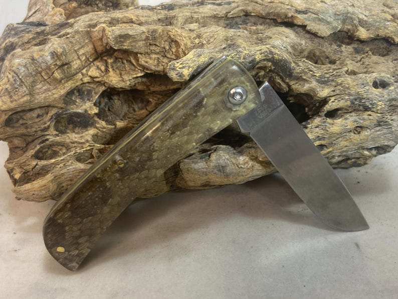 May include: A folding pocket knife with a partially open, stainless steel blade. The handle has a mottled green and brown pattern. The knife is resting on a piece of weathered wood.