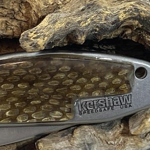 May include: A silver-toned folding knife handle featuring a textured, amber-colored honeycomb pattern inlay. The metal frame is engraved with the text Kershaw SPEEDSAFE USA and includes a small circular lanyard hole at the end of the handle.