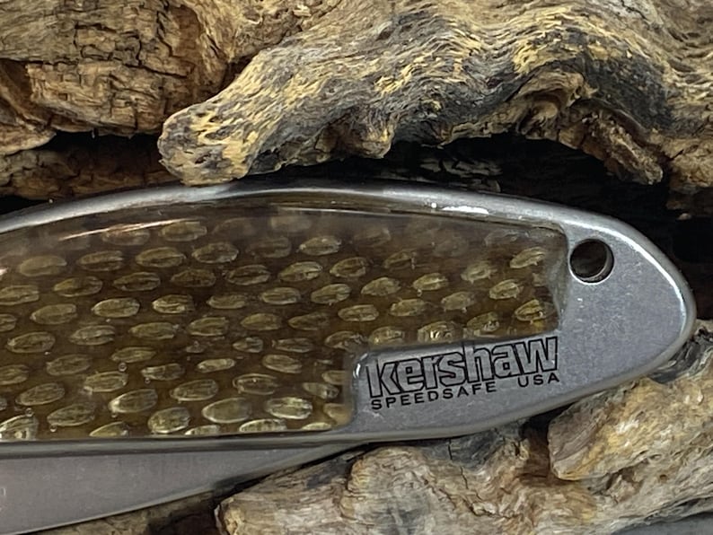 May include: A silver-toned folding knife handle featuring a textured, amber-colored honeycomb pattern inlay. The metal frame is engraved with the text Kershaw SPEEDSAFE USA and includes a small circular lanyard hole at the end of the handle.
