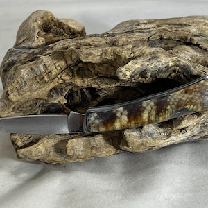 May include: A folding pocket knife with a metallic silver blade and a handle featuring a textured, mottled brown and cream pattern. The handle has a smooth, polished finish and a small circular hole at the end for a lanyard or attachment.