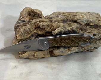 Könnte beinhalten: Ein Klappmesser mit einer silbernen Metallklinge und einer eingravierten Schlangengrafik. Der Griff hat eine strukturierte, honigwabenförmige braune Einlage und eine silberne Backe. Das Wort Kershaw ist am Ende des Griffs aufgedruckt.