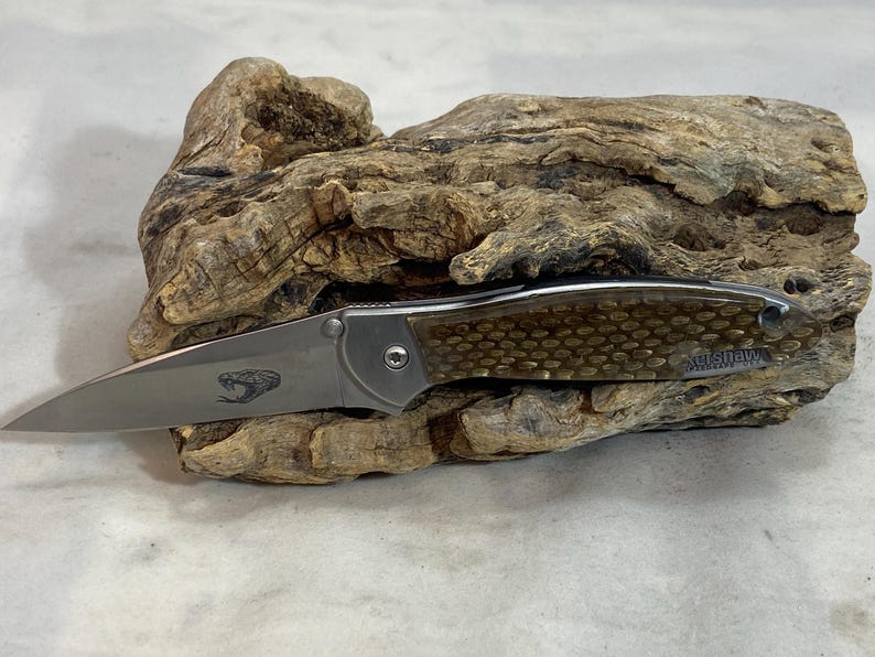 May include: A folding pocket knife with a silver metal blade featuring an etched snake graphic. The handle has a textured, honeycomb-patterned brown inlay and a silver bolster. The word Kershaw is printed on the handle near the end.