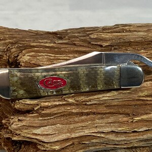 Pu&ograve; includere: Un coltello tascabile Case con un manico con motivo a pelle di serpente verde e marrone e una lama in acciaio inossidabile. Il coltello presenta un logo Case rosso e un apribottiglie. Il coltello &egrave; appoggiato su un pezzo di legno.