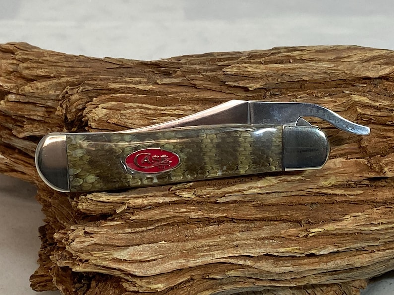 Pu&ograve; includere: Un coltello tascabile Case con un manico con motivo a pelle di serpente verde e marrone e una lama in acciaio inossidabile. Il coltello presenta un logo Case rosso e un apribottiglie. Il coltello &egrave; appoggiato su un pezzo di legno.