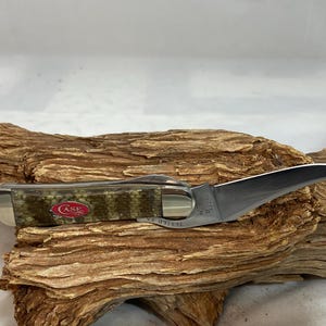 Pu&ograve; includere: Un coltello tascabile Case con una lama curva e un manico con motivo mimetico. Il coltello &egrave; aperto, mostrando l'affilata lama argentata. Il manico ha un logo Case rosso.