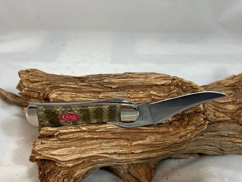 Pu&ograve; includere: Un coltello tascabile Case con una lama curva e un manico con motivo mimetico. Il coltello &egrave; aperto, mostrando l'affilata lama argentata. Il manico ha un logo Case rosso.