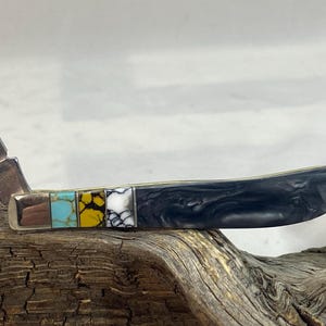 Puede incluir: Una navaja de bolsillo con una hoja de acero inoxidable y un mango con piedras decorativas incrustadas. El mango tiene un dise&ntilde;o veteado negro y una serie de incrustaciones de piedras de colores.