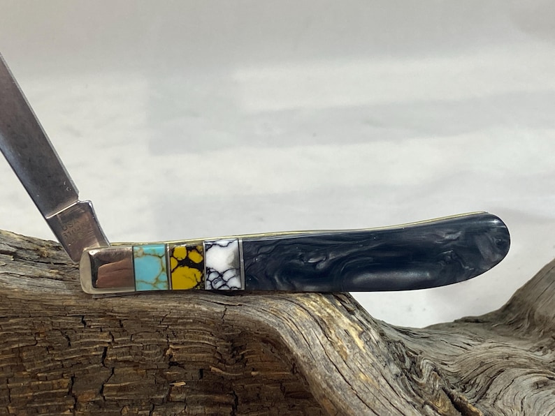 Puede incluir: Una navaja de bolsillo con una hoja de acero inoxidable y un mango con piedras decorativas incrustadas. El mango tiene un dise&ntilde;o veteado negro y una serie de incrustaciones de piedras de colores.