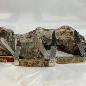 K&ouml;nnte beinhalten: Zwei klappbare Taschenmesser mit mehreren Klingen. Das linke Messer hat einen dunkelroten und schwarzen Griff, w&auml;hrend das rechte Messer einen strukturierten beigen Griff aufweist. Beide Messer haben silberne Metallbacken und ein Emblem.