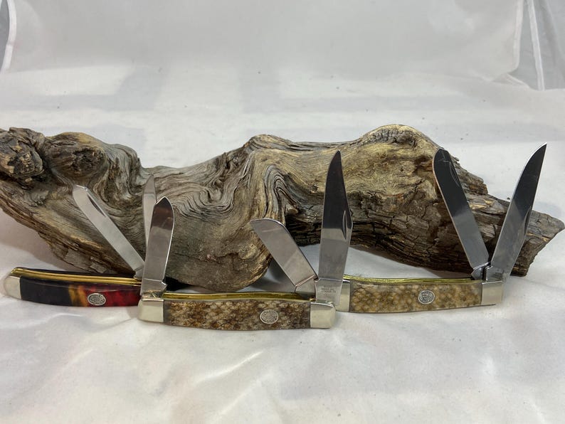 K&ouml;nnte beinhalten: Zwei klappbare Taschenmesser mit mehreren Klingen. Das linke Messer hat einen dunkelroten und schwarzen Griff, w&auml;hrend das rechte Messer einen strukturierten beigen Griff aufweist. Beide Messer haben silberne Metallbacken und ein Emblem.