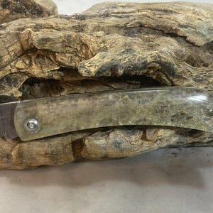 May include: A folding knife with a stainless steel blade and a handle made of a translucent, speckled material. The handle is secured with silver-colored rivets. The knife rests on a piece of weathered wood.