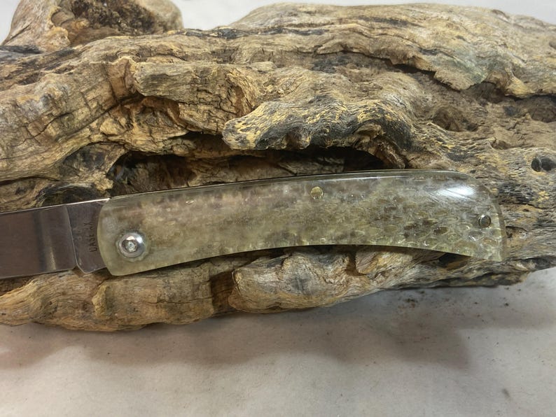 May include: A folding knife with a stainless steel blade and a handle made of a translucent, speckled material. The handle is secured with silver-colored rivets. The knife rests on a piece of weathered wood.