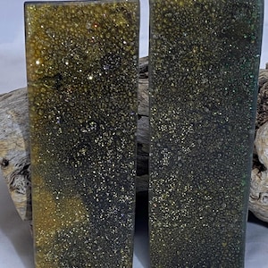 Könnte beinhalten: Zwei rechteckige Harzstücke mit gesprenkeltem, glitzerndem Aussehen. Das Harz ist hauptsächlich dunkelgrün und goldfarben, mit einer strukturierten Oberfläche. Die Stücke werden vor einem natürlichen Holzgrund gezeigt.