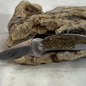 May include: A folding pocket knife with a silver metallic blade featuring a small snake engraving. The handle has a textured, golden-brown scale pattern and the text Kershaw SpeedSafe USA printed on the side near the lanyard hole.