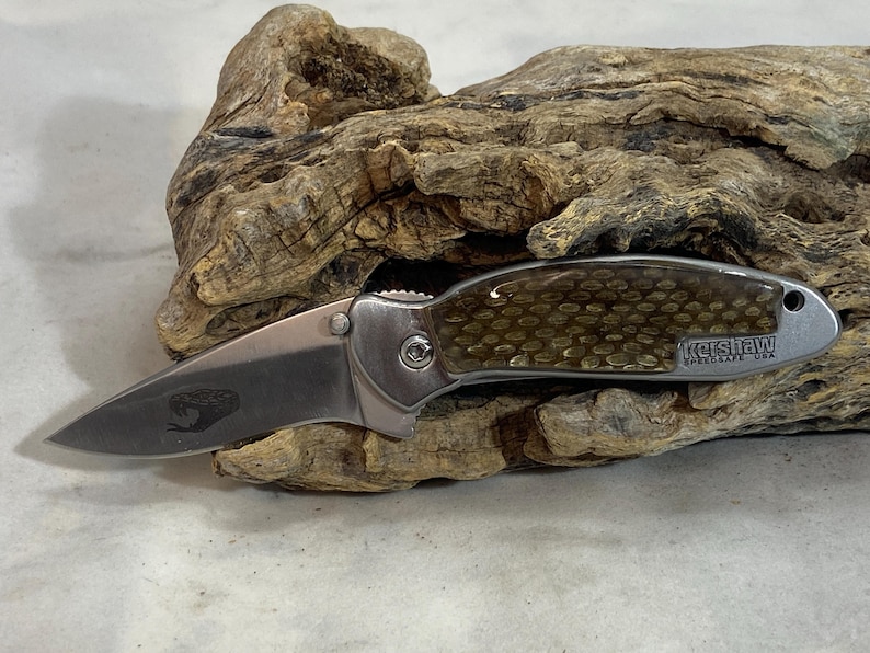 May include: A folding pocket knife with a silver metallic blade featuring a small snake engraving. The handle has a textured, golden-brown scale pattern and the text Kershaw SpeedSafe USA printed on the side near the lanyard hole.