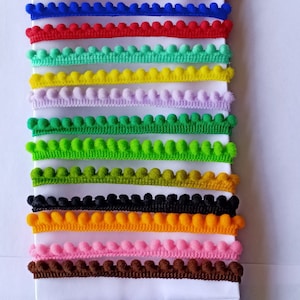 May include: Assortment of colourful pom-pom trim in various colours, including red, green, yellow, pink, blue, orange, brown, and white. The trim is arranged in rows on a white surface.