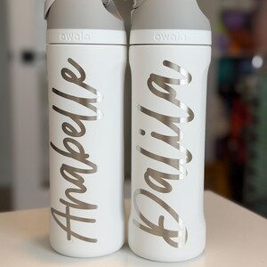 May include: Two white Owala water bottles with gray lids. One bottle has the name "Anabelle" in silver script, the other has "Dalila". The bottles are on a white surface.