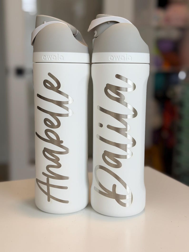 May include: Two white Owala water bottles with gray lids. One bottle has the name "Anabelle" in silver script, the other has "Dalila". The bottles are on a white surface.