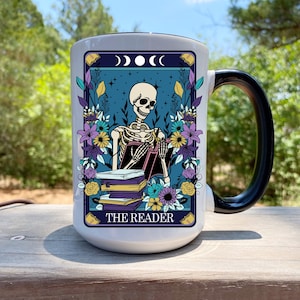 May include: White ceramic mug with a black handle featuring a tarot card design. The design includes a skeleton reading books, surrounded by flowers, with the text "THE READER" at the bottom. The background is a teal color with a crescent moon design.