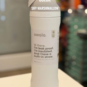 May include: A white Owala water bottle with a gray lid. The bottle has the text "Hi there. I'm leak proof. I'm insulated. And I have a built-in straw." The bottle is 24 oz (710 ml). The color is "Shy Marshmallow."
