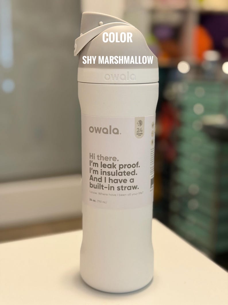 May include: A white Owala water bottle with a gray lid. The bottle has the text "Hi there. I'm leak proof. I'm insulated. And I have a built-in straw." The bottle is 24 oz (710 ml). The color is "Shy Marshmallow."