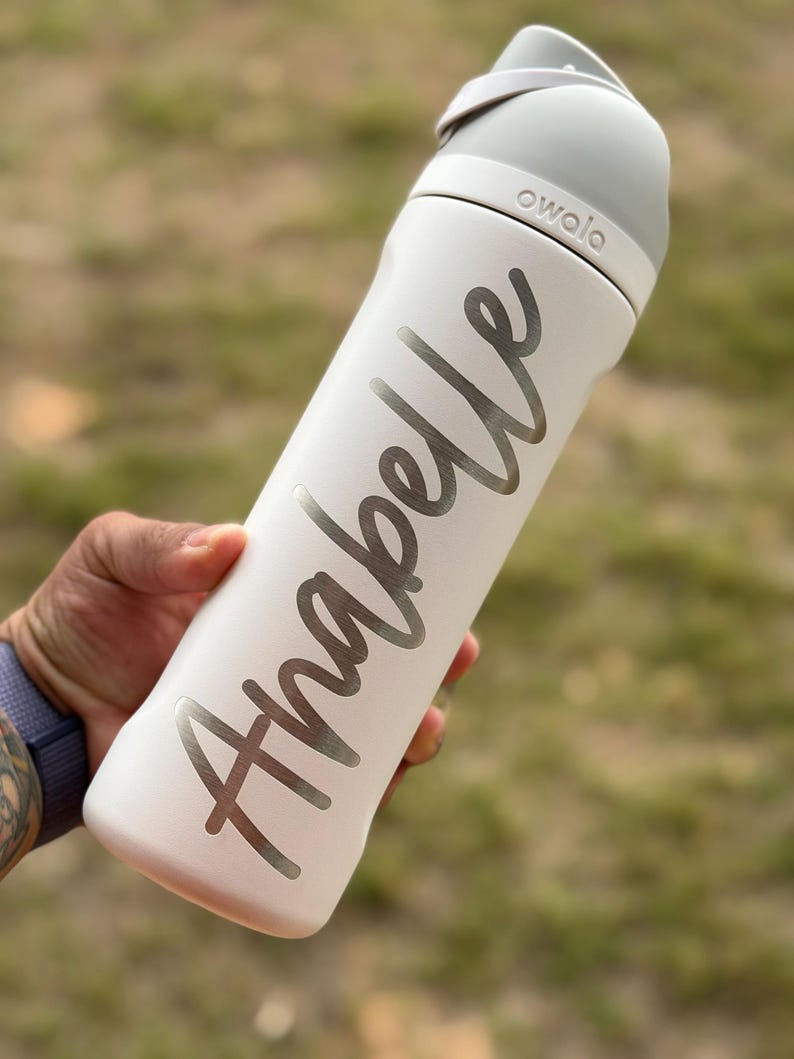 May include: A white Owala water bottle with a gray lid and handle. The name "Anabelle" is written in silver script on the bottle. The bottle is held against a blurred green and brown background.