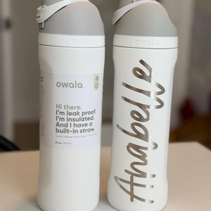 May include: Two white Owala water bottles with gray lids and accents. One bottle has a label with text, the other has the name "Anabelle" in silver script. Each bottle is 24 oz (710 mL) and has a built-in straw.