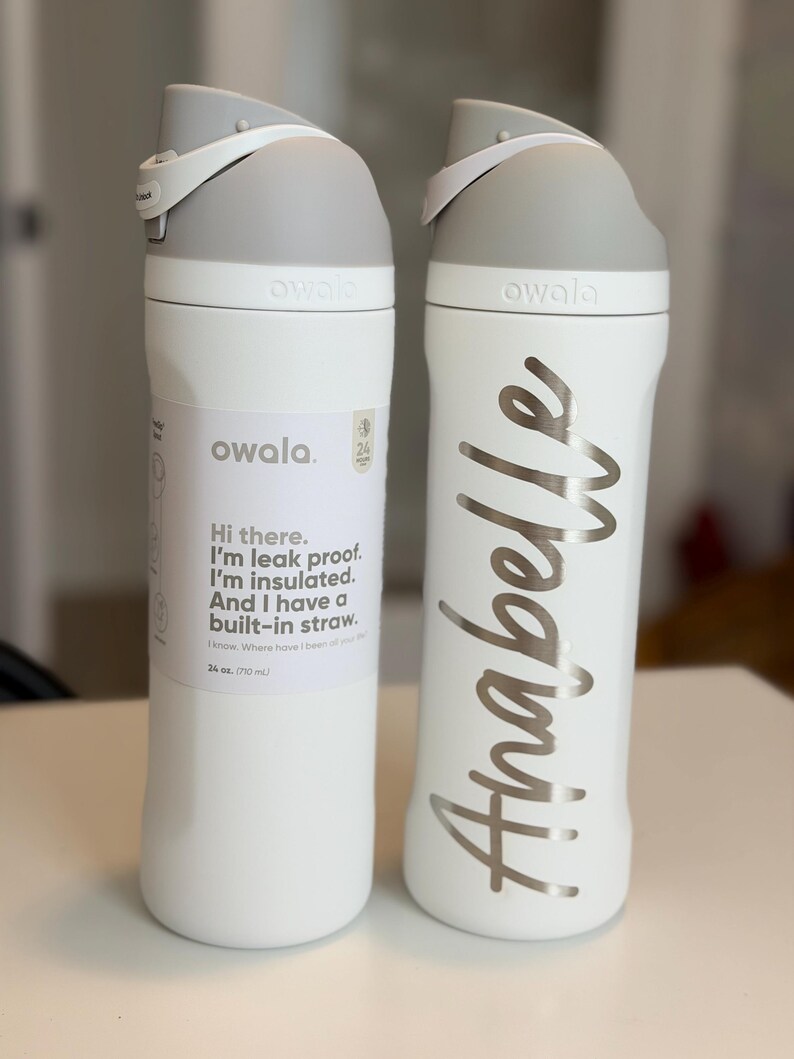 May include: Two white Owala water bottles with gray lids and accents. One bottle has a label with text, the other has the name "Anabelle" in silver script. Each bottle is 24 oz (710 mL) and has a built-in straw.