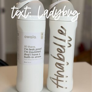 May include: Two white Owala insulated water bottles. One bottle has the name "Anabelle" in silver script. The other bottle has text that reads "Hi there. I'm leak proof. I'm insulated. And I have a built-in straw." The bottles are 24 oz (710 mL).